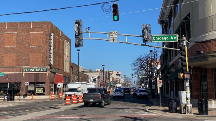 Downtown Naperville street with buildings, traffic lights and cars in view, as well as orange traffic barrels where streetscape work will happen