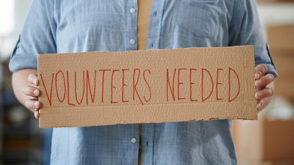 A person holds a sign that says "Volunteers Needed"