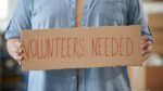 A person holds a sign that says "Volunteers Needed"
