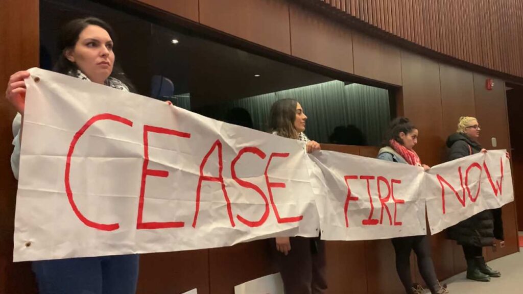 Protestors in the Naperville Municipal Center's council chambers holding a banner that reads "cease-fire, now"