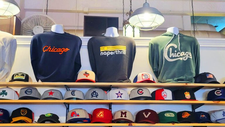 Three sweatshirts on display, the one in the middle says "Naperthrill" and the other two say "Chicago" baseball hats are on display underneath.