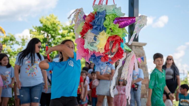 A young girl in a blue t-shirt swings at a multicolored pinata.
