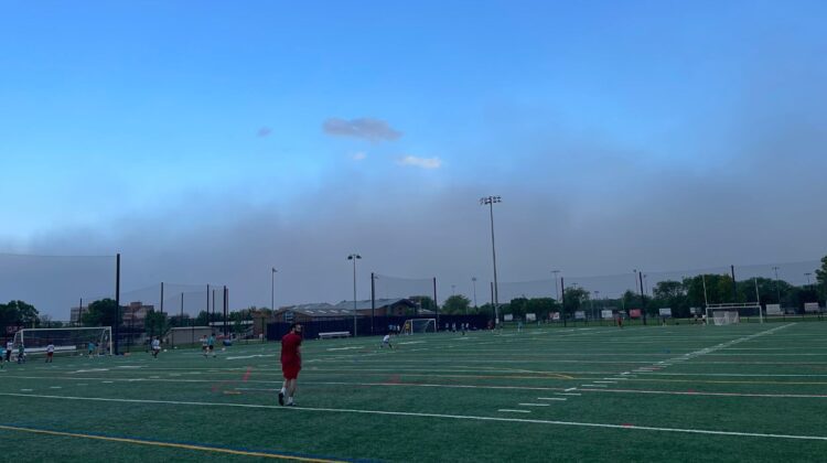 Man in red outfit walks across soccer field with large dust storm cloud visible in distance