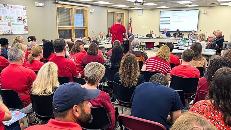 Wide shot of Naperville School District 203 board meeting with board in front of room and audience members in red shirts visible with speaker at podium